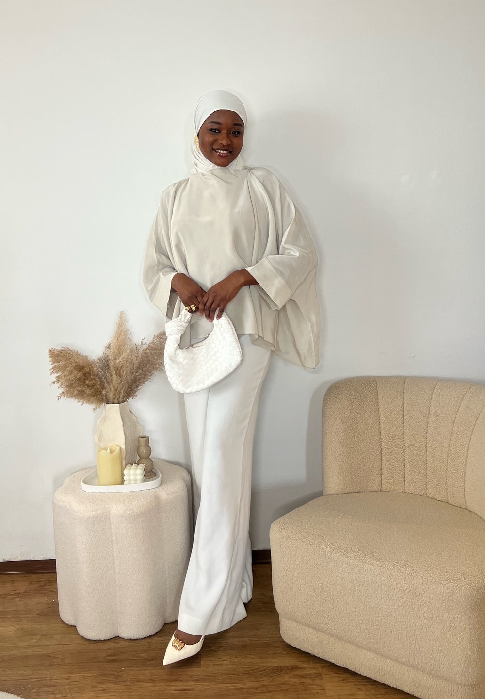 Bluză albă vaporoasă cu mâneci largi, combinată cu pantaloni albi mulați. Accesorizată cu o geantă texturată albă și pantofi ascuțiți. Mobilier bej deschis în fundal.