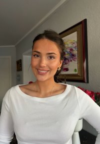 Jeune femme aux cheveux foncés attachés en arrière, souriante, portant un haut blanc et des boucles d'oreilles dorées en créole, debout à l'intérieur avec des œuvres encadrées et des fleurs derrière elle.