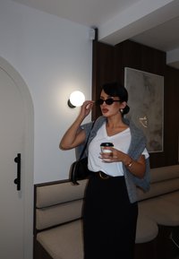 Femme portant des lunettes de soleil, tenant une tasse de café, vêtue d'une chemise blanche, d'une jupe noire, et d'un pull gris posé sur les épaules dans un espace moderne de café.