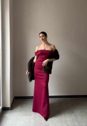 Mujer con un vestido largo burdeos sin hombros, sosteniendo una estola de piel oscura, de pie en una habitación minimalista con una gran ventana.