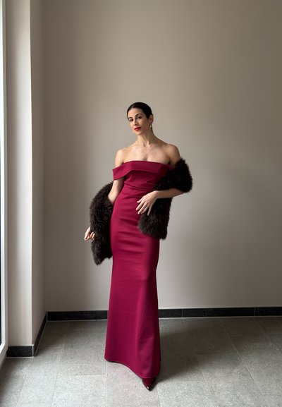 Mujer con un vestido largo burdeos sin hombros, sosteniendo una estola de piel oscura, de pie en una habitación minimalista con una gran ventana.