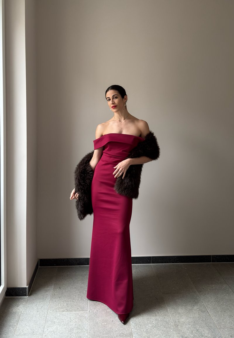 Γυναίκα με μακρύ βελούδινο φορέμα χωρίς ώμους σε μπορντό χρώμα, κρατώντας ένα σκοτεινό γούνινο κασκόλ, στέκεται σε έναν λιτό χώρο με μεγάλο παράθυρο.