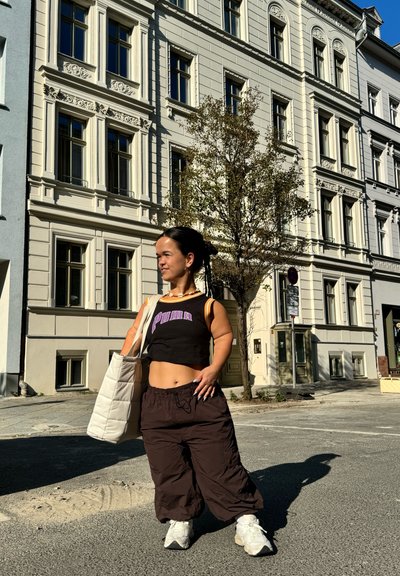 Top corto negro con logo de "PUMA", pantalones baggy marrones, zapatillas blancas y una bolsa de tote acolchada de color claro. Fondo urbano.