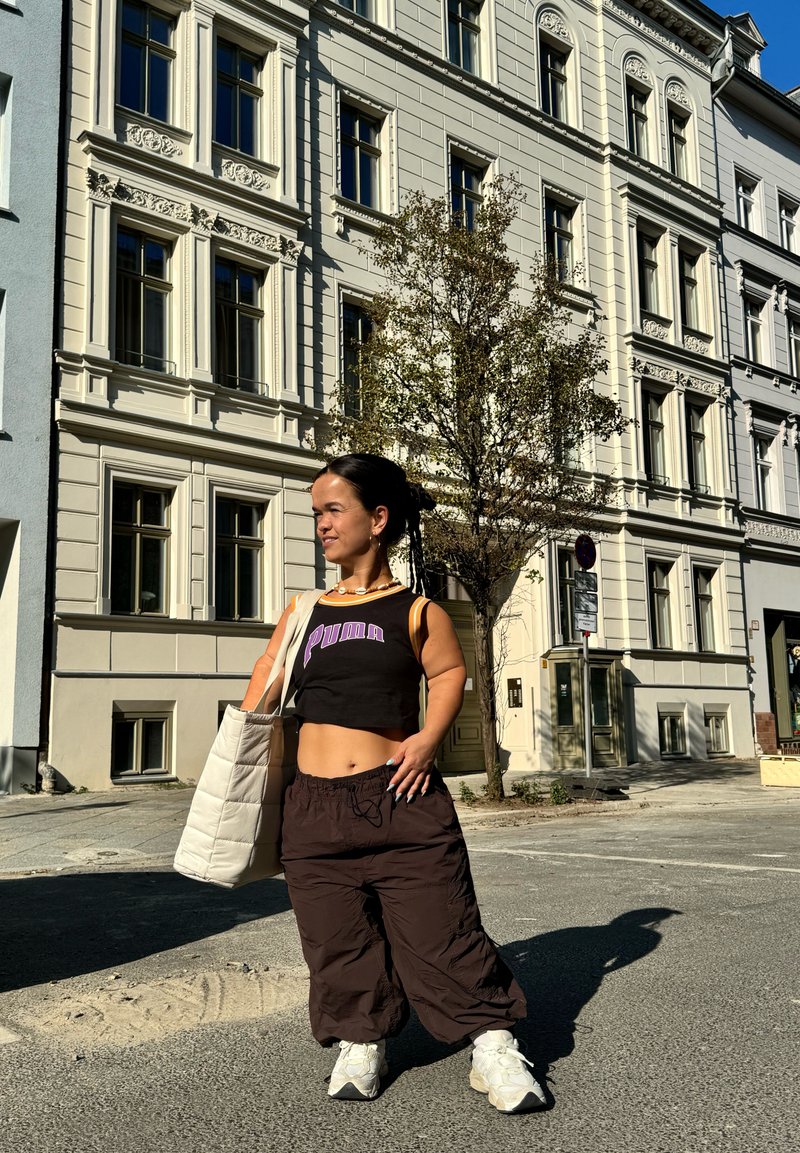Μαύρο κοντό αμάνικο μπλουζάκι με το λογότυπο "PUMA", καφέ φαρδιά παντελόνια, λευκά αθλητικά παπούτσια και μία ελαφριά κουβέρτα τσάντα. Αστική σκηνή στο φόντο.