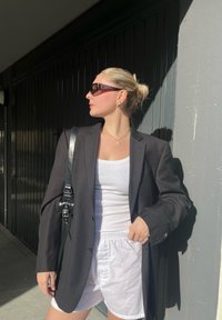 Μαύρο σακάκι από ύφασμα πάνω από λευκή μπλούζα και λευκά σορτς. Αξεσουάρ περιλαμβάνουν ένα πράσινο κολιέ με pendant και γυαλιά ηλίου. Ορατή υφή στην επιφάνεια.