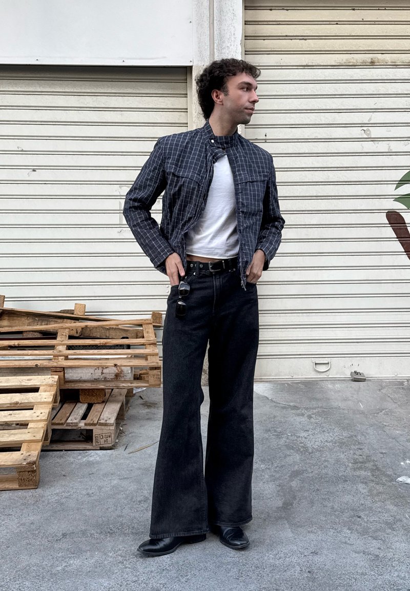Blue checked jacket, white t-shirt, dark wide-leg jeans, black belt, and black shoes. The background features wooden pallets and a plant.