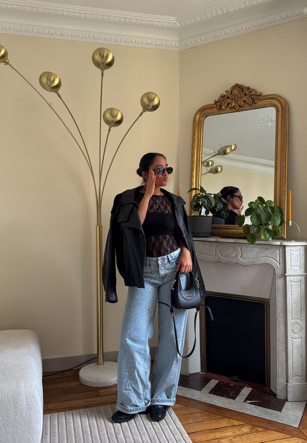 Mujer con top de encaje negro, chaqueta de cuero, vaqueros brillantes y gafas de sol, de pie junto a una chimenea con plantas y un espejo con marco dorado en una sala de estar.