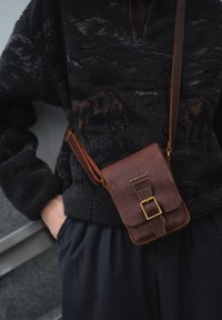 Sac bandoulière en cuir marron à surface texturée, fermeture à boucle et sangle réglable. Porté sur un pull en polaire noir.
