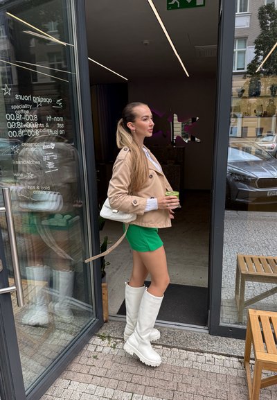Abrigo trench beige, pantalones cortos verdes y botas altas blancas. Sosteniendo una bebida, de pie en una entrada con paneles de vidrio y asientos exteriores visibles.