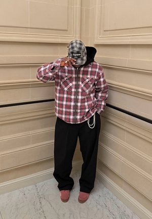 Persona con chaqueta de cuadros rosa, pantalones negros, zapatos rosas y una gorra gris de Supreme, de pie contra paredes beige con paneles, con una mano ajustando la gorra.