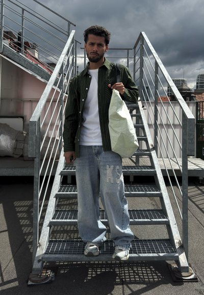 Camisa de rayas verdes sobre una camiseta blanca, jeans holgados azules claros con un logo de K, sosteniendo una bolsa verde arrugada, de pie en escaleras de metal.