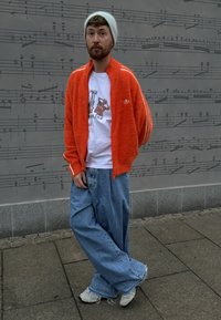 Orange fuzzy zip-up jacket with white stripes, worn over a white T-shirt and loose blue jeans. Grey beanie and white trainers complete the outfit.