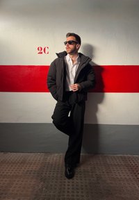 Homme portant une veste noire, une chemise blanche et des lunettes de soleil, appuyé contre un mur avec une bande rouge horizontale et le marquage "2C" dans une zone de stationnement.