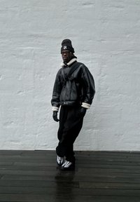 Giacca di pelle nera con colletto in shearling, guanti neri e pantaloni neri larghi. Scarpe sportive bianche con dettagli argento. Sfondo neutro.