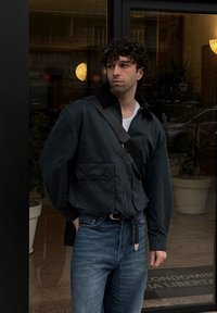 Jeune homme portant une veste noire, un jean bleu, des lunettes de soleil sur la tête, et portant un sac à bandoulière noir, debout près de portes vitrées avec des plantes à l'intérieur.