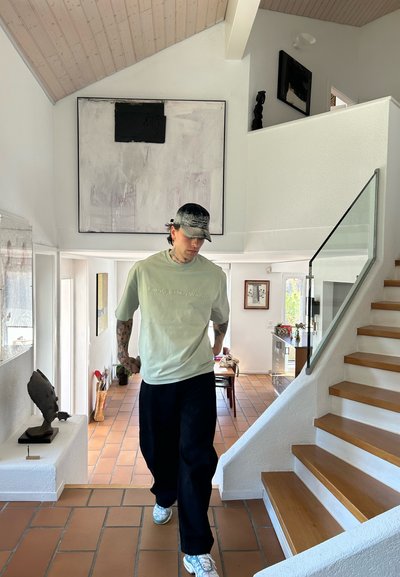 Camiseta de algodón verde claro con texto en relieve, pantalones sueltos negros y zapatillas multicolores. Interior moderno con escaleras de madera y obras de arte.
