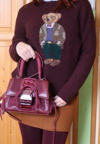 Άτομο που φοράει μπορντό πουλόβερ με σχέδιο αρκούδας σε σακάκι, κρατώντας μια τσάντα Steve Madden σε ταιριαστή μπορντό απόχρωση με κορδόνια και διάτρητα πάνελ.