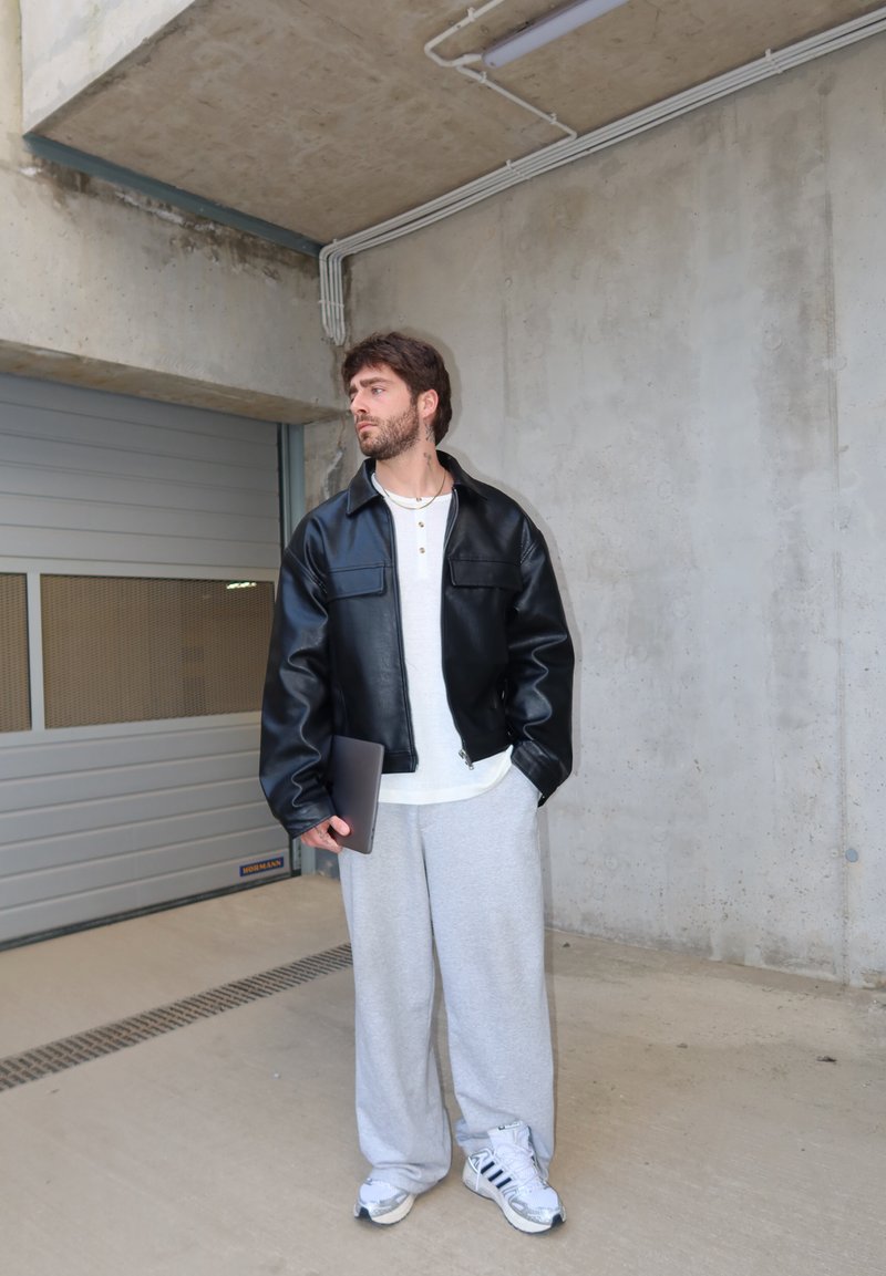 Uomo con giacca di pelle nera e pantaloni della tuta grigi che tiene un laptop, in piedi in un parcheggio coperto in cemento vicino a una porta del garage chiusa.