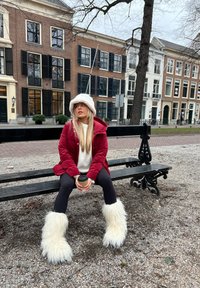 Veste matelassée rouge, chapeau fluffy blanc, pull crème, leggings noirs et bottes en fourrure blanche. Assise sur un banc noir dans un cadre urbain.