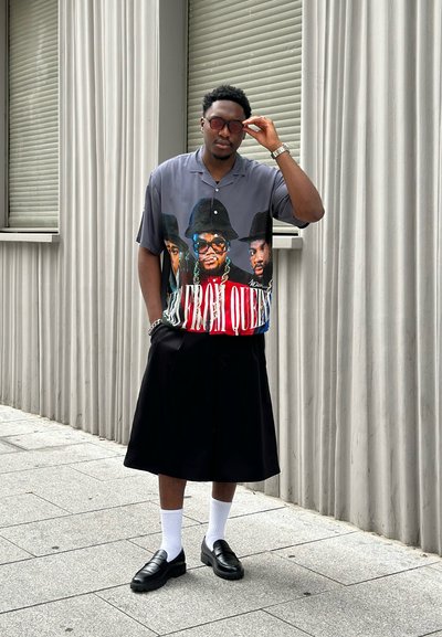 Camisa gris de manga corta con estampado gráfico, falda negra hasta la rodilla, mocasines negros y calcetines blancos. De pie contra una pared de rayas verticales.