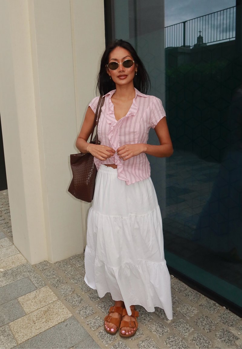 Femme portant des lunettes de soleil rondes, un chemisier à manches courtes rayé rose, une longue jupe blanche, des sandales brunes et un grand sac cabas brun foncé.