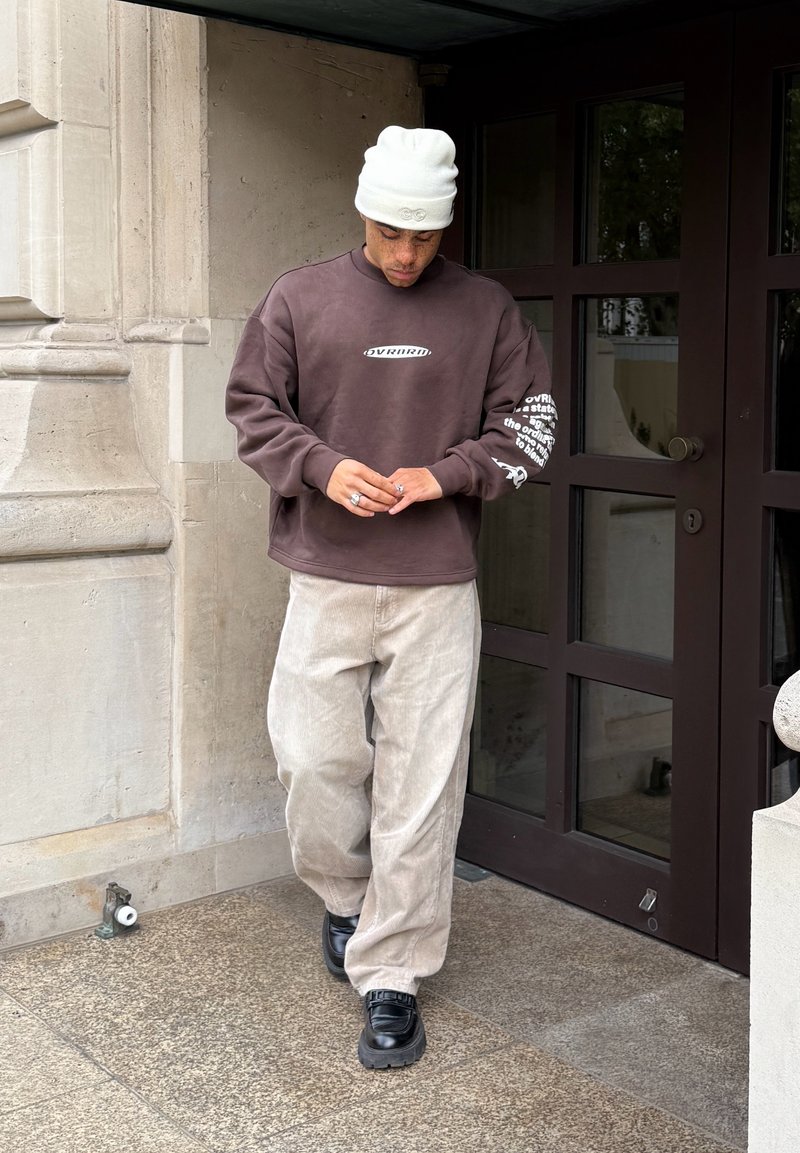 Muž nosiaci bielu čiapku, hnedú mikinu, béžové kordové nohavice a čierne topánky, kráčajúci po kamennej podlahe pri vchode do budovy.