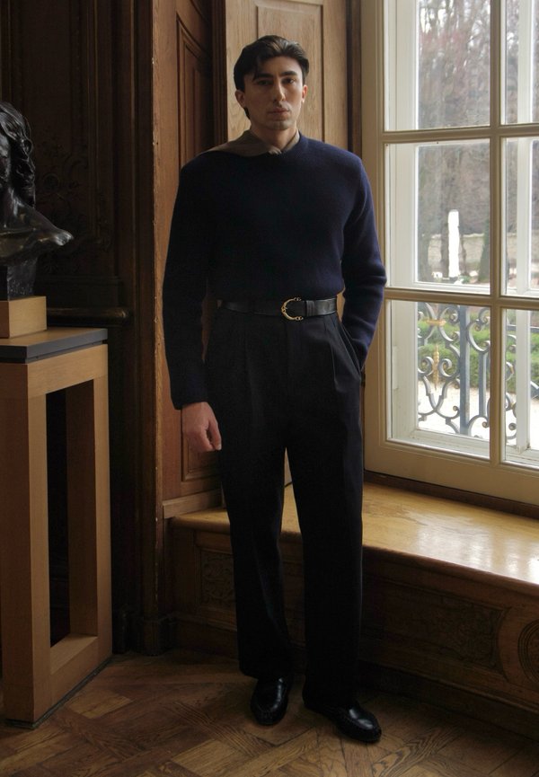 Man in zwarte broek, marineblauwe trui en zwarte schoenen die binnen staat bij een groot raam en een houten paneel met een bronzen buste op een tafel.