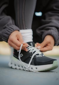 Ember szürke melegítőnadrágot viselve fűzi be a fehér cipőfűzőket egy fekete-fehér futócipőn, amelynek talpán látható párnázás, szabadban.