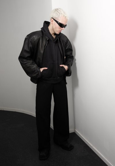 Persona con cabello rubio corto, con gafas de sol negras, chaqueta de cuero negra sobre sudadera negra con capucha, pantalones negros, de pie contra una pared blanca.