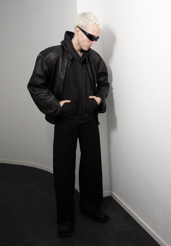 Person mit kurzem blondem Haar, die schwarze Sonnenbrille trägt, schwarze Lederjacke über schwarzem Hoodie, schwarze Hose, steht vor einer weißen Wand.