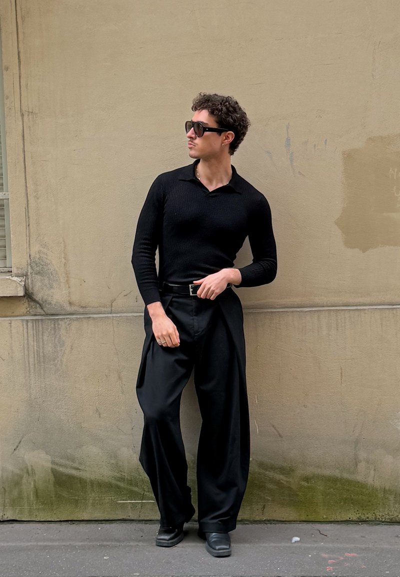 Άνδρας φοράει μαύρο μακρυμάνικο πουκάμισο, φαρδιά μαύρα παντελόνια, ζώνη και γυαλιά ηλίου, στέκεται σε μπεζ τοίχο, κοιτώντας στο πλάι με το ένα χέρι στη μέση.