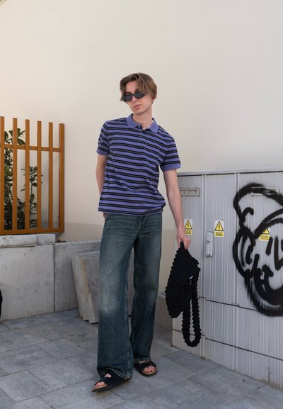 Camisa de polo morada a rayas, pantalones vaqueros de denim holgados, sandalias negras, sosteniendo un bolso negro con textura. El fondo presenta una pared beige y una cerca marrón.