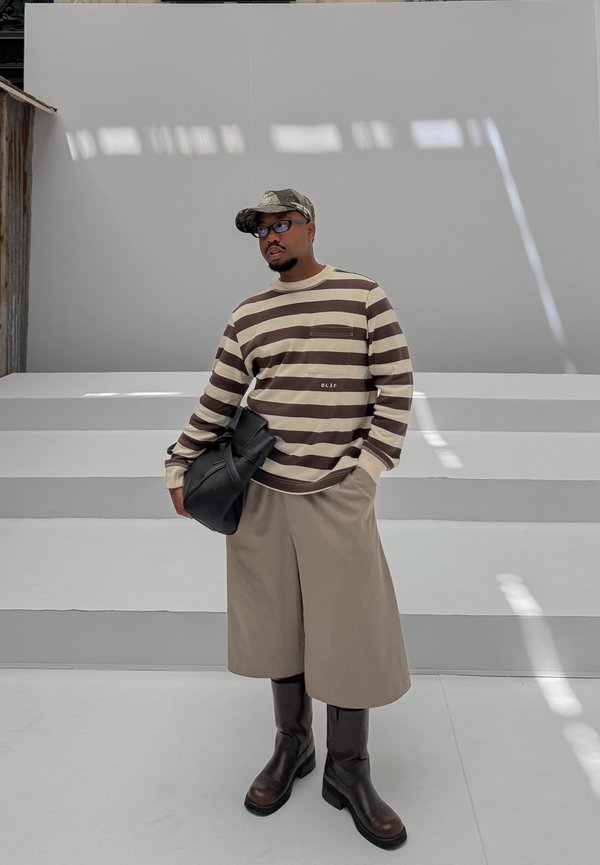 Mand iført en brun og beige stribet langærmet skjorte, beige vide shorts, høje brune støvler, camouflage kasket og briller, der holder en sort taske.