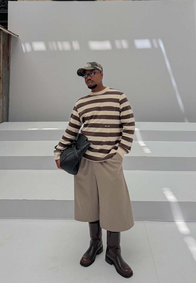 Homme portant une chemise à manches longues rayée marron et beige, un short large beige, de grandes bottes marron, une casquette camouflage et des lunettes, tenant un sac noir.