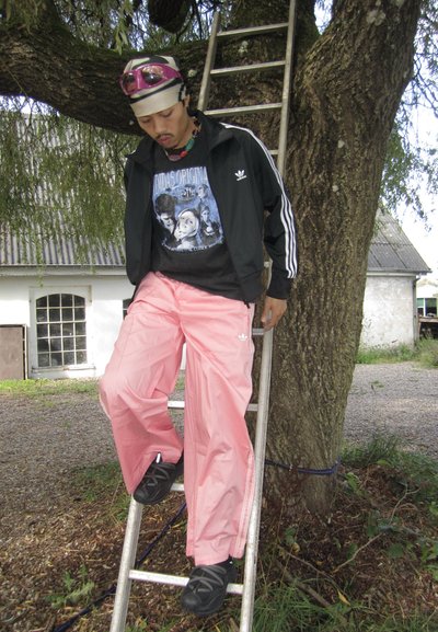 Atleta vistiendo una chaqueta de chándal negra de Adidas, camiseta gráfica y pantalones de chándal rosas, de pie en una escalera apoyada contra un árbol, con gafas de sol en la cabeza.