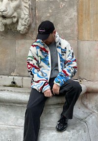 Veste en polaire avec un motif abstrait multicolore, base blanche, dotée d'une fermeture éclair, portée sur un pull gris et un jean noir.