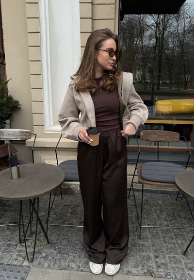 Chaqueta oversized de mezcla de lana gris claro, blusa ajustada marrón, pantalones de satén anchos marrón oscuro, zapatillas blancas, sosteniendo un vaso de café para llevar.