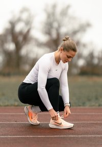 Vitt tröja med långa ärmar, svarta leggings och ljusa sneakers med orangeröda detaljer. Person som knyter skosnören på en löparbana.