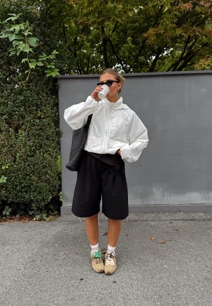 Chaqueta blanca con cremallera y cuello alto; pantalones cortos negros de corte holgado; bolsa de tote negra; zapatillas grises; sosteniendo una taza de café; gafas de sol.