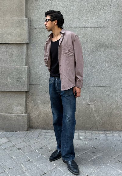 Hombre con gafas de sol, camiseta sin mangas negra, camisa suelta color malva con botones, vaqueros azules y zapatos negros, de pie en una acera de la ciudad mirando hacia la izquierda.