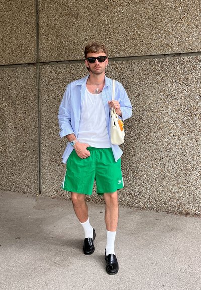 Camiseta sin mangas blanca, camisa de botones a rayas azules, pantalones cortos verdes con rayas blancas, mocasines negros y calcetines blancos. Sosteniendo una bolsa ligera.