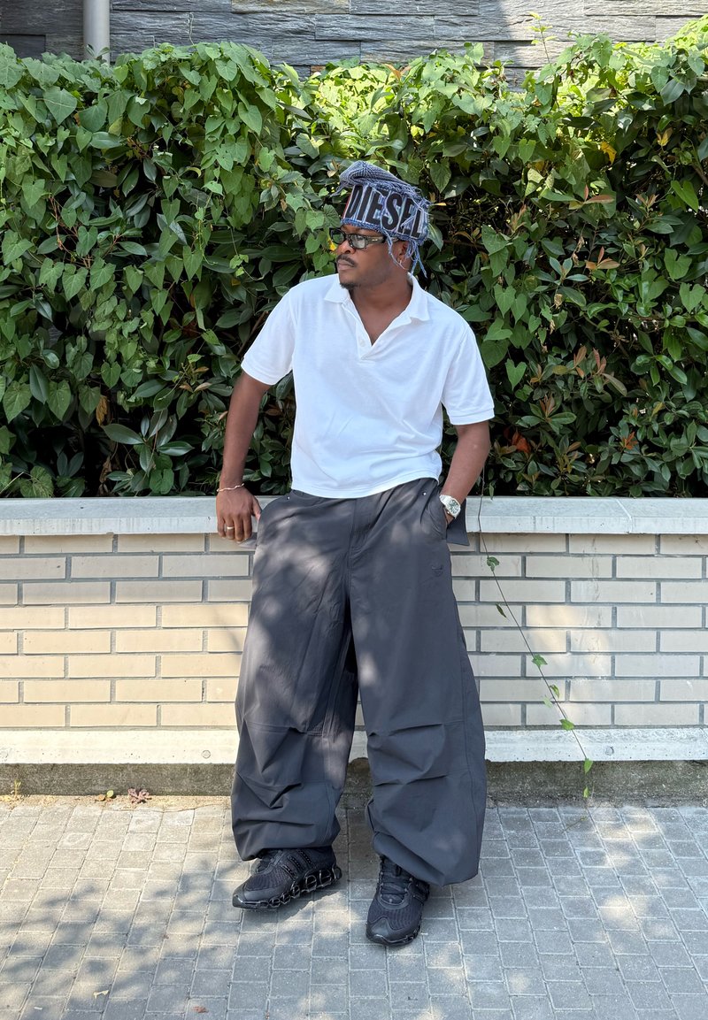 Chemise polo blanche, pantalon gris foncé oversized à la coupe ample, baskets noires, lunettes de soleil et chapeau à motifs. Feuillage vert en arrière-plan.