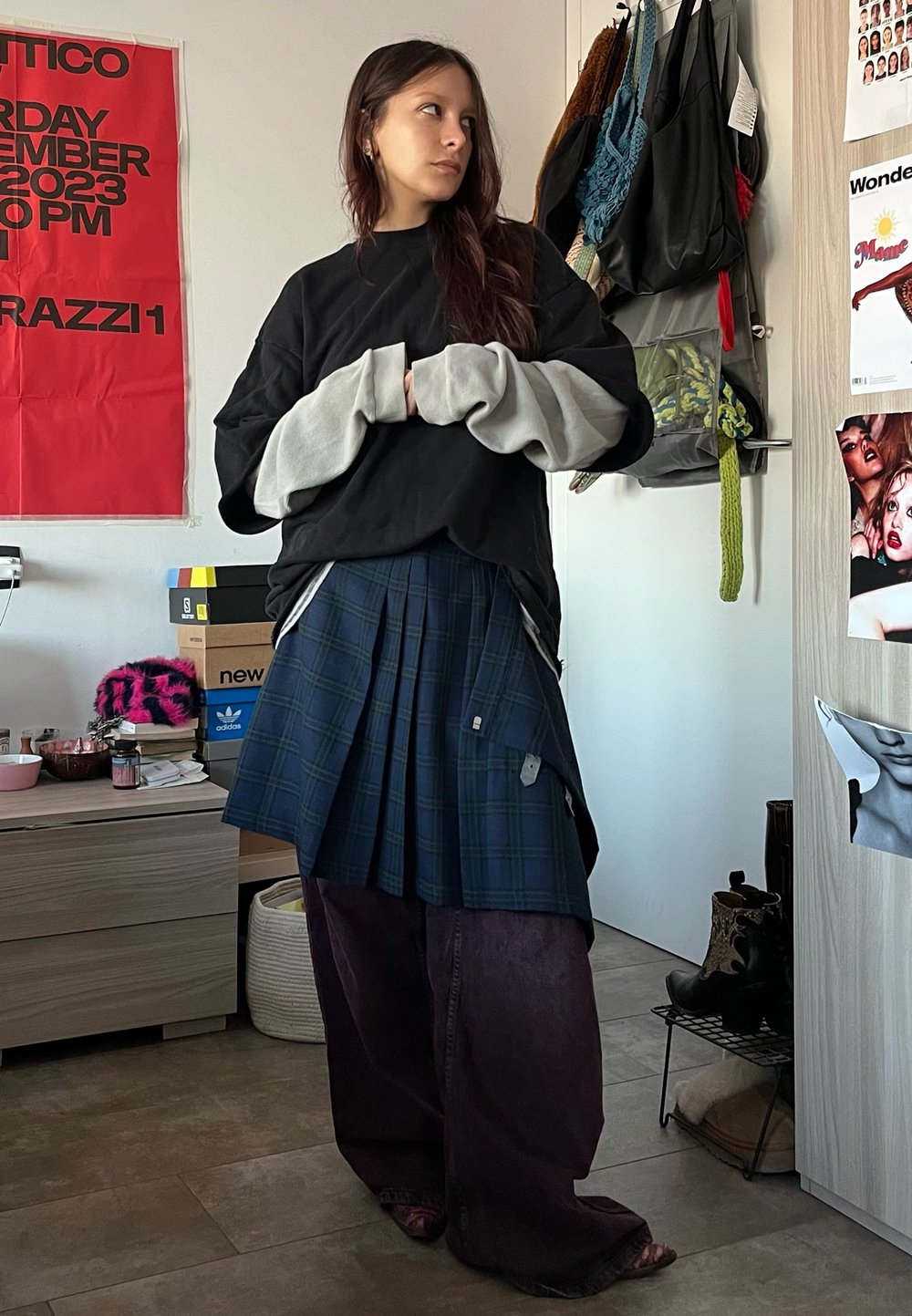 Svart oversize sweatshirt med grå detaljer, blå rutigt plisserad kjol och lösa lila kordbyxor. Inomhusmiljö med olika föremål synliga.