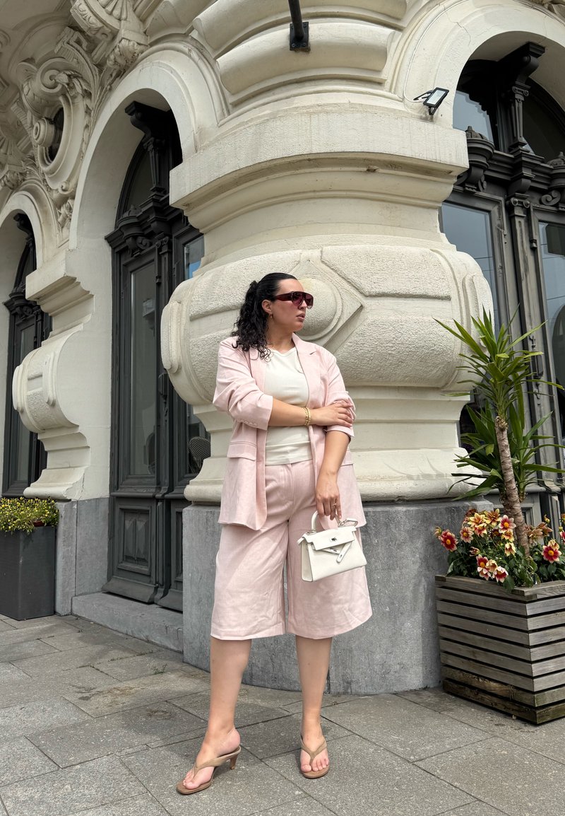 Femme en tailleur rose pâle et sandales, debout tenant un sac à main blanc près d’un bâtiment en pierre orné, avec de grandes fenêtres et des jardinières.