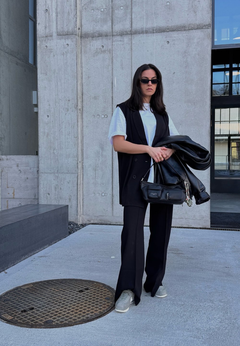 Colete preto sobre uma t-shirt branca, calças pretas e sapatos cinza. Segurando uma bolsa preta e uma jaqueta de couro. Fundo de concreto com uma grelha redonda.