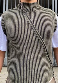 Pletený bezrukávový svetr s vertikálními černými a zelenými pruhy, vysokým límcem a diagonálním zipem na přední straně. Měkká textura.
