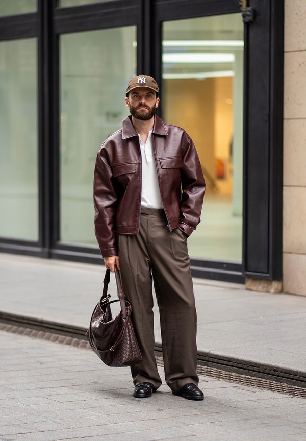 Un hombre barbudo con una gorra marrón, una chaqueta de cuero burdeos y pantalones holgados está de pie frente a un edificio con fachada de cristal y sostiene un bolso grande.