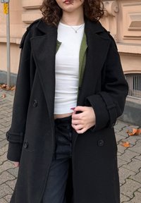 Manteau noir en laine, croisé, avec de gros boutons, de longueur moyenne, larges revers et manches retroussées, porté sur un haut blanc et un cardigan vert.