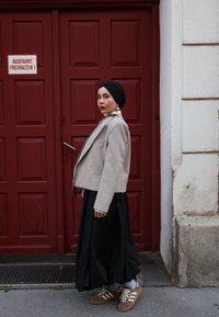 Blazer court gris clair avec une poche unique, superposé sur une longue jupe noire. Porté avec des baskets beiges et accessoirisé avec de grandes boucles d'oreilles.