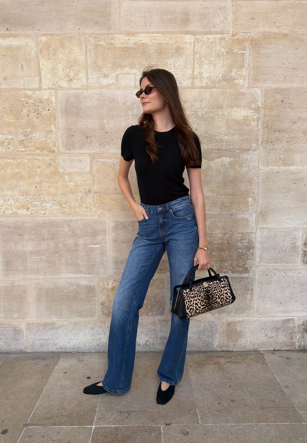 Mujer con top negro, vaqueros azules, zapatos negros y gafas de sol sosteniendo un bolso de estampado de leopardo mientras está de pie junto a una pared de piedra.